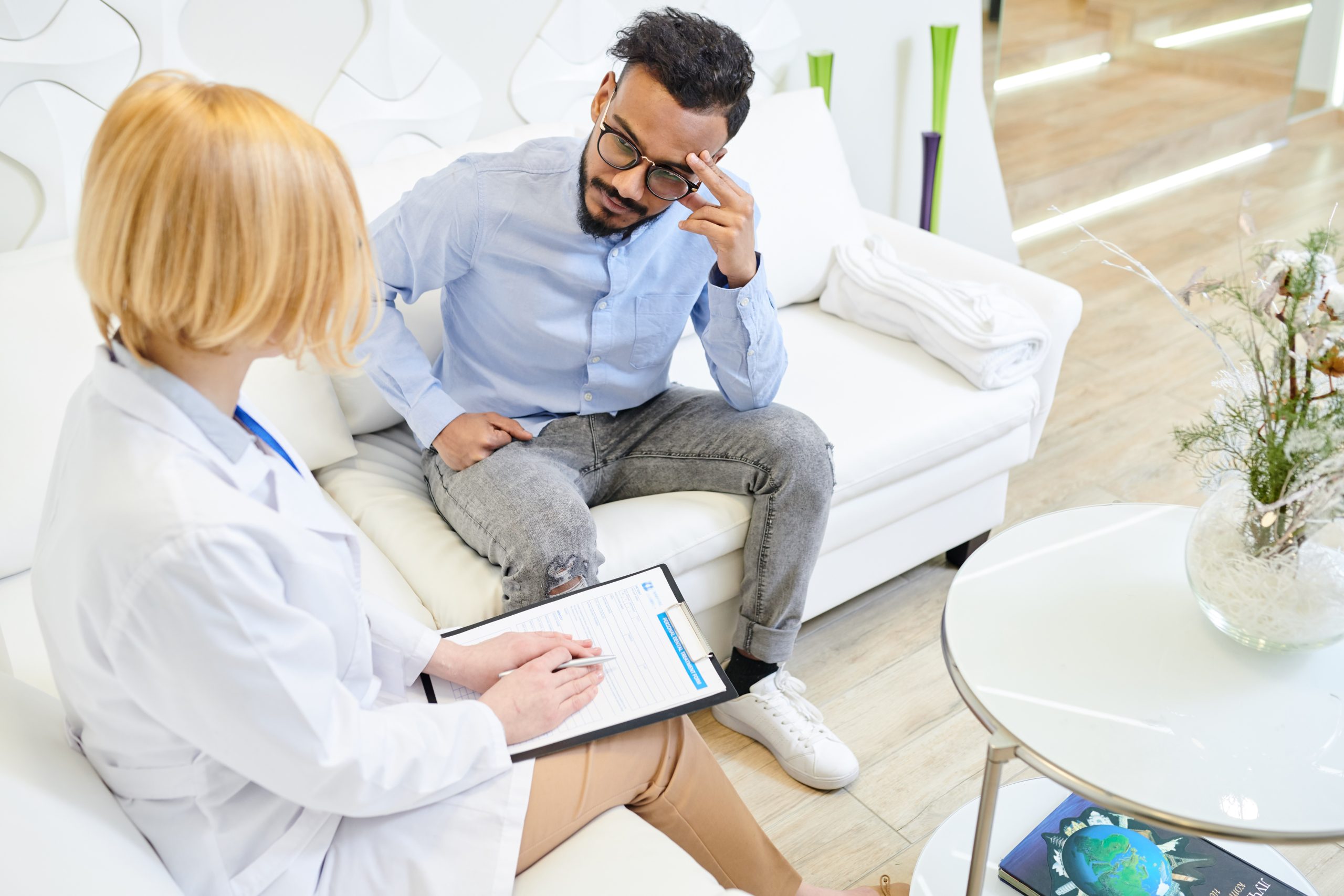 Highly professional practitioner having appointment: handsome young patient sitting on sofa and listing symptoms while she listening to him with concentration